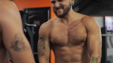 Genç adam spor salonunda çalışıyor. Ciddi şekilde odaklanmış. Aynanın önünde çıplak vücut kaslarını gösteriyor.