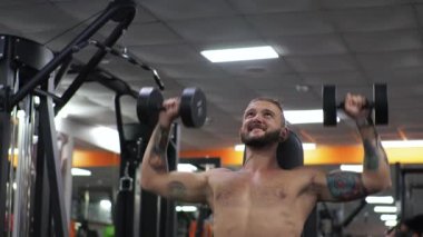 Genç adam spor salonunda çalışıyor. Odaklanıyor. Kollarını sallıyor, halteri itiyor.