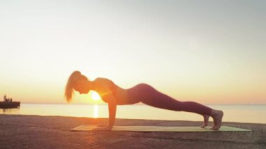Kumsalda yoga yapan genç sarışın kadın güneşin doğuşunun ya da batışının arka planında