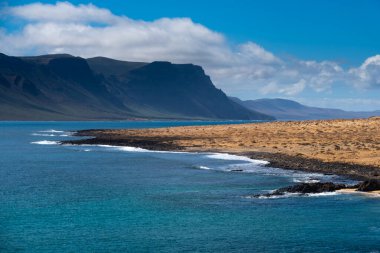 La Graciosa 'nın engebeli volkanik kıyı şeridinde siyah kayalarla karşılaşan turkuaz suları bulunur. Mavi gökyüzünün altındaki görkemli uçurumlar yükseliyor. Ziyaretçiler kumlu yolda yürüyor