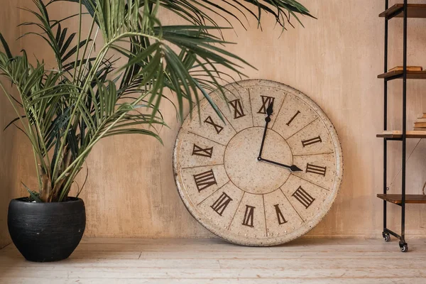 A large French provincial style clock with Roman dial stands on the floor in a beige interior next to a floor planter with a plant and a bookshelf. Loft style. New year. Christmas Eve.