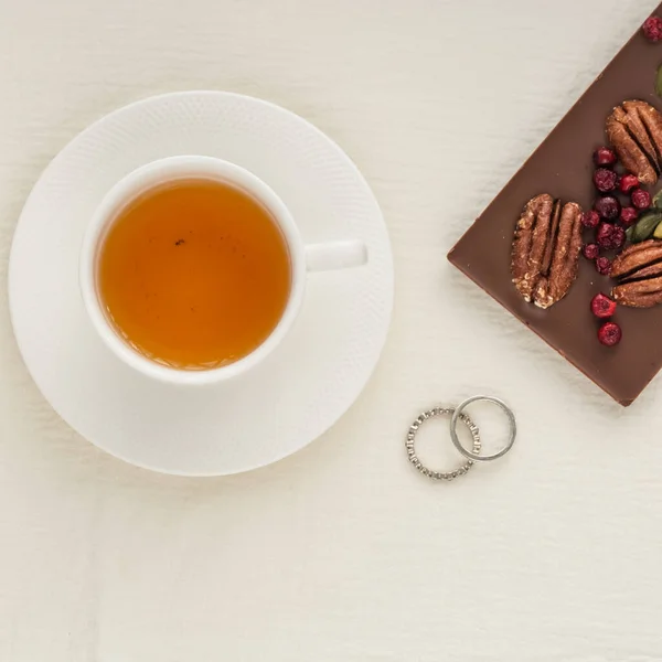 Chocolate bar decorated with nuts and berries on a white background tablecloth and mugs, close-up, top view. Flat lay. Nuts pecans and pistachios in delicious desserts. Morning tea party concept.
