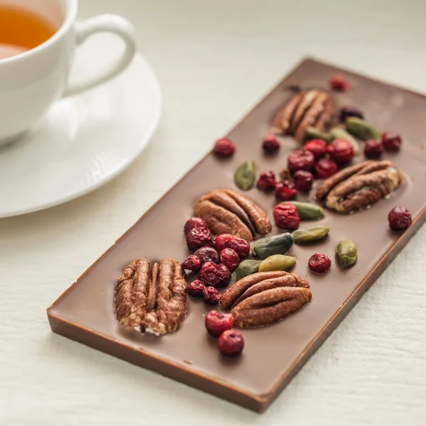 Chocolate bar decorated with nuts and berries on a white background tablecloth and mugs, close-up. Nuts pecans and pistachios in delicious desserts. Morning tea party concept.