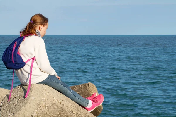 10代の女の子は海の近くの大きな石の上に座ってヘッドフォンで聞きます — ストック写真