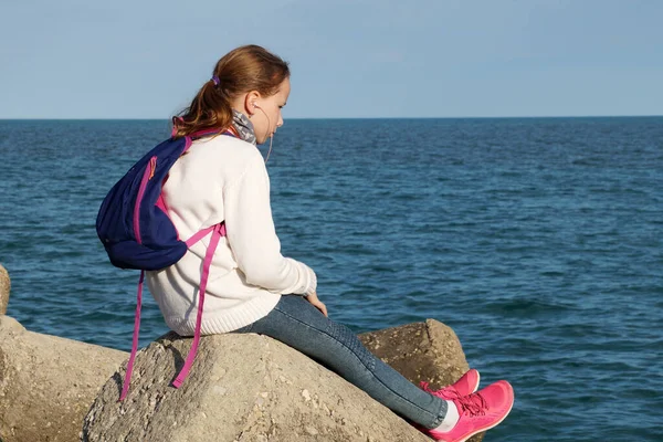 10代の女の子は海の近くの大きな石の上に座ってヘッドフォンで聞きます — ストック写真