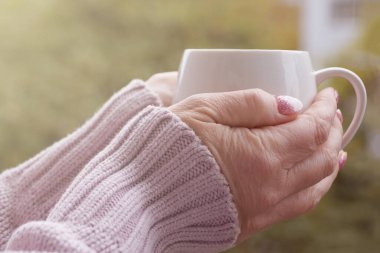 Sıcak kazak giymiş kadın elleri güneşli bir günde arka planda sıcak bir fincan tutar.
