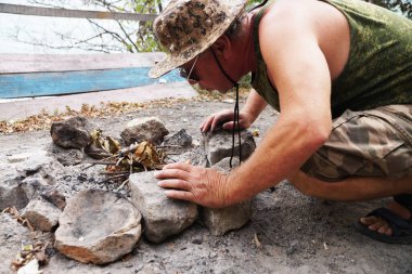 Şapkalı bir adam doğada şenlik ateşi yakıyor..