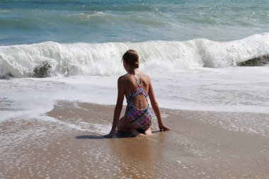 Genç kız ıslak kumda tek başına oturuyor ve denize ve beyaz dalgalara bakıyor.