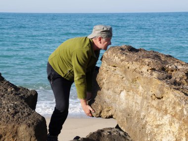 Bir adam deniz kıyısında büyük bir taşı kaldırır..