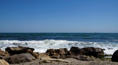 Güneşli bir günde dalgalı ve mavi gökyüzü olan kayalık bir sahil.