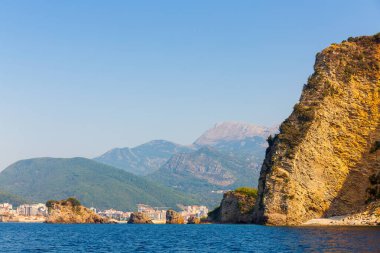 Kayalar, dağlar, güneşli sahil ve gök mavisi deniz.