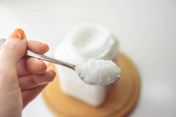 Female hand holds a spoon with white sea salt. Blurred background of jar and wooden board on white table.