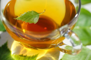 Huş yaprağıyla birlikte cam bardakta yeşil çay. Bardağın etrafında beyaz bir masada içkiyle duruyor..