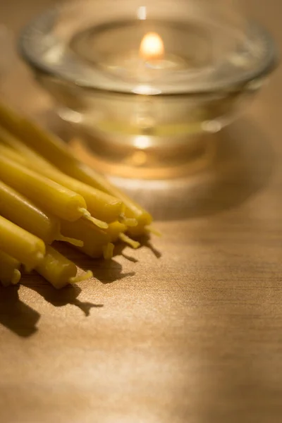 Heap of new long candles on the table. One low candle burns in a glass round candlestick in blurred background.