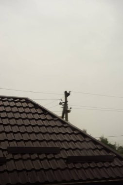 Yağmurlu bulutlu bir günde elektrik direğinin yanında kahverengi kiremitli çatılı ev