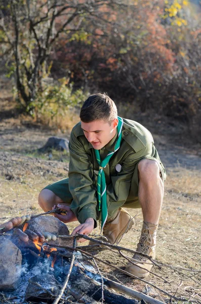 Rapaz escuteiro cozinhando salsichas em paus sobre fogueira — Fotografia de Stock