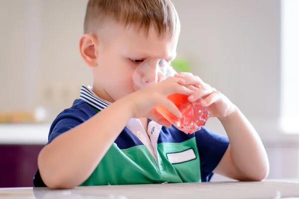 Jongen op keuken tafel drinken glaasje rode SAP — Stockfoto