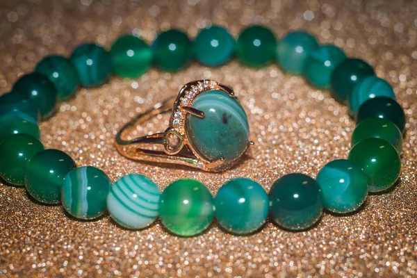 Modern gold ring and beaded bracelet made from green striped agate on glitter background.