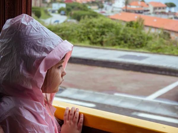 Girl in a pink raincoat looking out a window on a rainy summer day in Cantabria