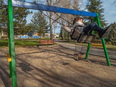 Güneşli ve soğuk bir kış gününde şehir parkında salıncakta sallanan ceketli ve çizmeli bir kız.