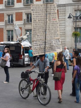 Madrid, İspanya; 23 Mayıs 2011. 15-M protestoları sırasında Puerta del Sol 'daki insanlar arasında bisiklet üzerinde protesto pankartı gösteren yaşlı bir adam. Fotoğraf 23 Mayıs 2011 'de Madrid, İspanya' da çekildi.