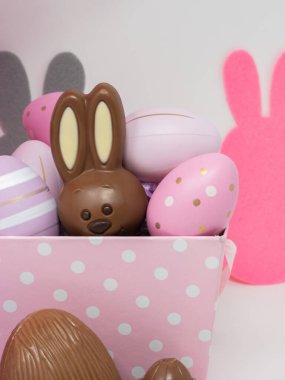 Çikolatalı Paskalya tavşanı pembe sepetin içinde dekoratif yumurtaların arasına saklanmış, beyaz arka planda tavşan siluetleri olan.