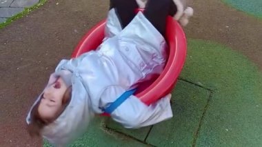 Gümüş ceketli ve atkılı küçük kız kış günü renkli bir parkta dönüyor.