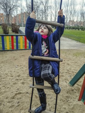Paltolu ve eşarplı ilkokul kızı bulutlu bir kış gününde oyun parkında merdivene tırmanıyor.