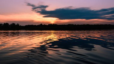 Manzara, gölün üzerinde gün batımı. Kıyı şeridinin silueti ve bulutlu gökyüzü