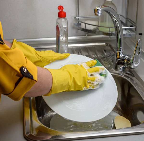 A woman in rubber gloves washes dishes in the kitchen sink, hands only