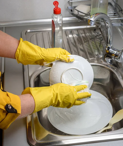 A woman in rubber gloves washes dishes in the kitchen sink, hands only