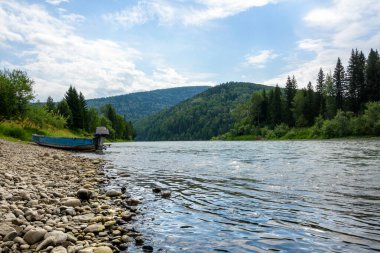 Gündüz manzarası bir dağ nehri ile kozalaklı ormanların arasında kayalık bir sahilde motorlu tekne