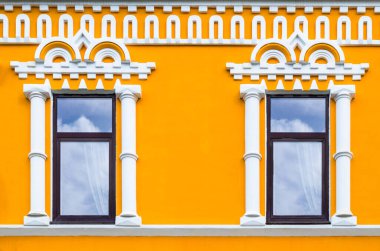 Sarı cephede iki büyük pencere. Gökyüzünün yansıması ve pencerelerde bulutlar. Duvardaki süs.