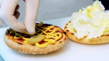 Lezzetli hamburgerin detaylı hazırlığı. Şefin eli hamburgerin üzerine salatalık parçaları koyuyor. Fast food restoranında çizburger yapmak. Yakın plan..