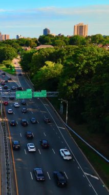 Çıkış işaretleri ve ağaçlar ve binalar arasındaki kontrastla dolu bir otoyol. New York 'un Queens ilçesinde yol boyunca giden arabaların hava manzarası. Dikey video.