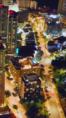 Honolulu, Oahu 'da gece görüşü. Aydınlatılmış sokaklar, yüksek binalar, çatı havuzu ve palmiye çizgili caddeler canlı bir kentsel atmosfer yaratıyor. Dikey video.