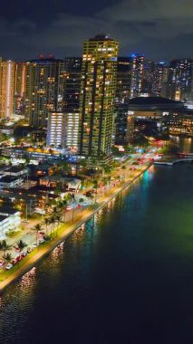 Honolulu, Oahu 'da gece görüşü. Rıhtıma bakan binalar, sokak lambaları ve palmiye ağaçları okyanusa yansıyarak dinamik bir kıyı manzarası yaratıyor. Dikey video.
