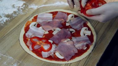 Eldivenli eller pizzaya pastırma, mantar ve domates soslu kırmızı dolmalık biber ekliyor. Ahşap yüzeyde un..