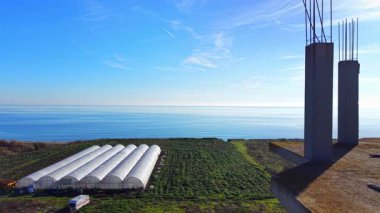 Yeşil alanda, kıyı şeridi yakınlarında bitmemiş beton kolonları olan beyaz seralar. Çok katlı bir binanın inşaatı. Hava görünümü.