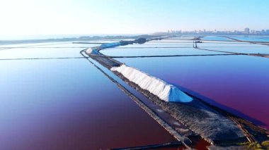 Günbatımında tuz üretim bölgesinin geniş bir manzarası. Büyük beyaz tuz yığınları şehir siluetine ve denize doğru giden yollar..