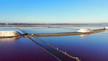 Tuz havuzlarındaki geleneksel ahşap yapıların havadan görünüşü. Sakin su mavi gökyüzünü ve endüstriyel ufku yansıtır. Hazırlanmış beyaz tuz yığını
