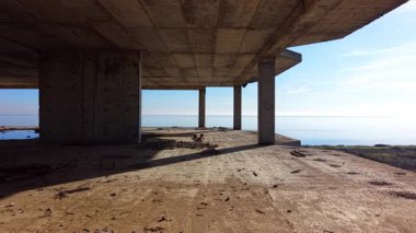 Bitmemiş beton bir yapının içinden yüksek açılı bir görüntü. Zemin toz ve enkaz ile kaplı. Deniz ve açık mavi gökyüzü sütunlardan görülebilir.