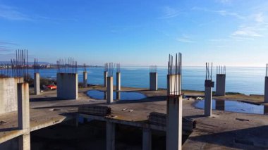 Geniş bir beton çatı manzarası ve sayısız dikey sütunlar. Su birikintileri mavi gökyüzünü yansıtır. Arka planda deniz ve kıyı görünüyor. Hava görünümü.