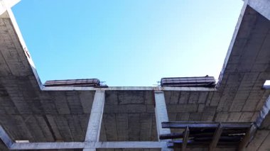 Bir binanın tamamlanmamış beton yapısına bakıyorum. Görünür kirişler, zemin döşemeleri ve açık mavi gökyüzüne karşı ahşap şekiller. Hava görünümü.
