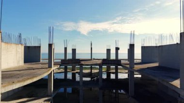 Gün doğumunda denize bakan beton sütunları ve yerdeki su birikintilerini gösteren bitmemiş bir binanın geniş açılı görüntüsü. Hava görünümü.