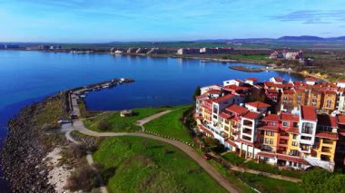 Kıyı şeridi boyunca kırmızı kiremitli çatıları olan çok katlı binaların geniş hava görüntüsü, eğri bir taş iskele ve mavi gökyüzünün altında sakin bir koy. Bulgaristan.