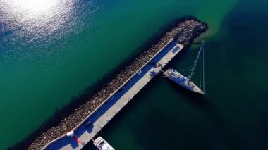 Açık yeşil deniz suyunda beton ve taş bir dalgakırana demirlenmiş büyük bir yelkenlinin hava görüntüsü. Taş iskeleye yanaşmış yelkenlinin üst görüntüsü.