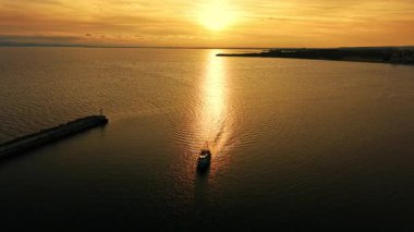 Deniz yüzeyindeki altın yansıma boyunca hareket eden tek bir teknenin havadan görüntüsü. Bir iskele silueti ve parlak bir güneşin altında bir kıyı şeridi..