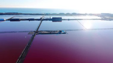 Pembe suyla geometrik tuzlukların geniş açılı görüntüsü. Su yüzeyinde parlak güneş yansıması. Endüstriyel binalar ve deniz arka planda görülebilir. Açık ufuk.
