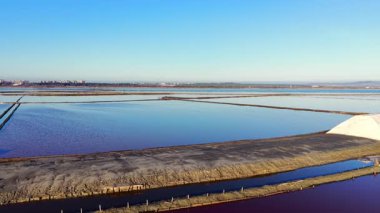 Hava görüntüsü, beyaz tuz yığınıyla endüstriyel toprak seti. Göletlerde hala mavi ve pembe su var. Ufuktaki uzak şehir ve tepeler.